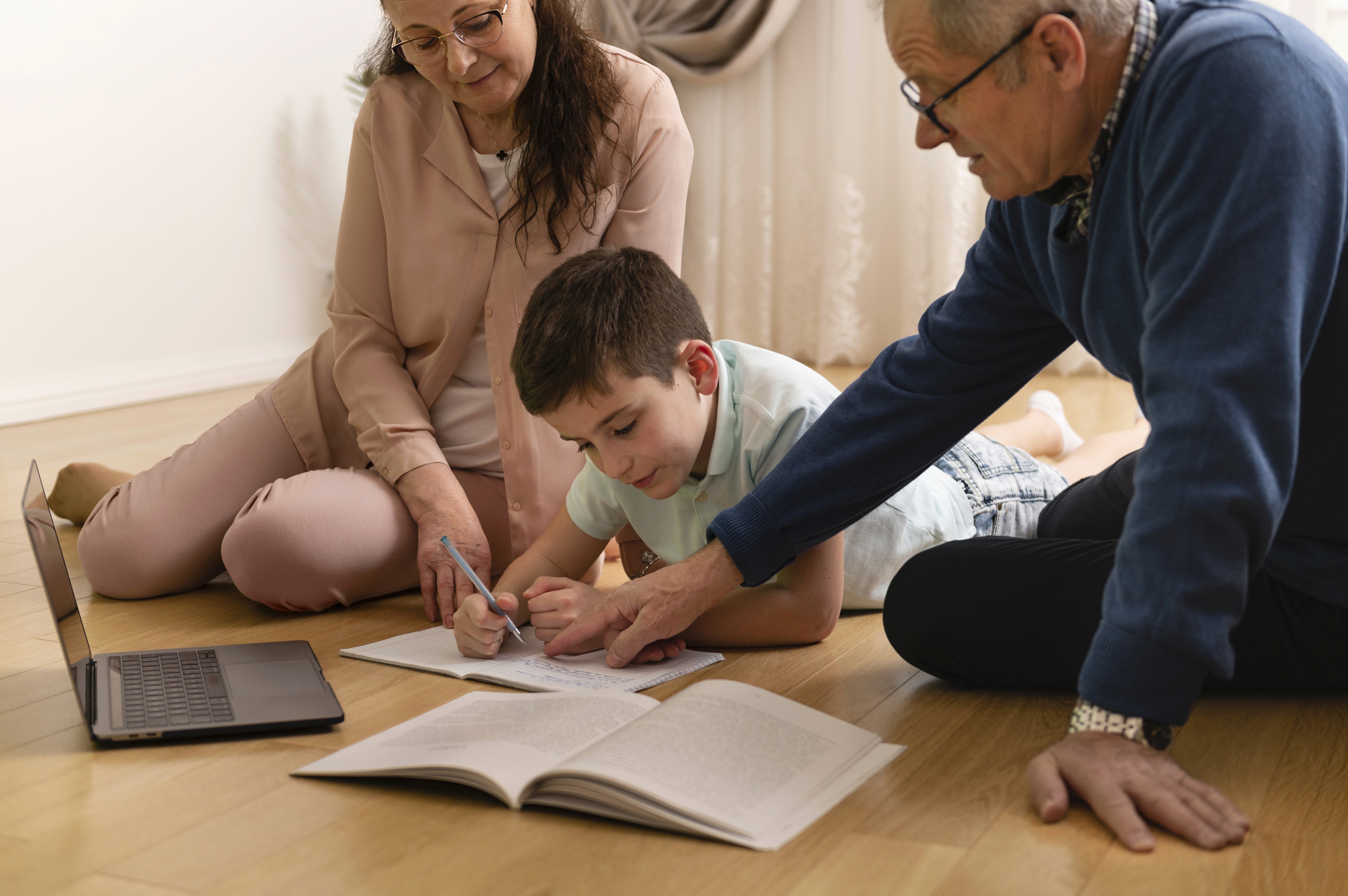 criança com livros, deitada no chão e escrevendo. Dois adultos estão ao seu lado, ajudando.