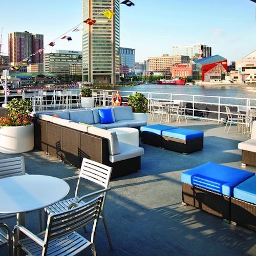 Outdoor rooftop seating area with sofas and tables overlooks a city waterfront with boats, tall buildings, and blue sky.