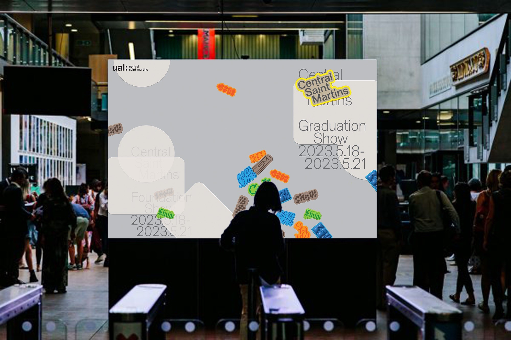 Person sitting in front of a digital screen displaying Central Saint Martins Graduation Show details with people walking in the background.