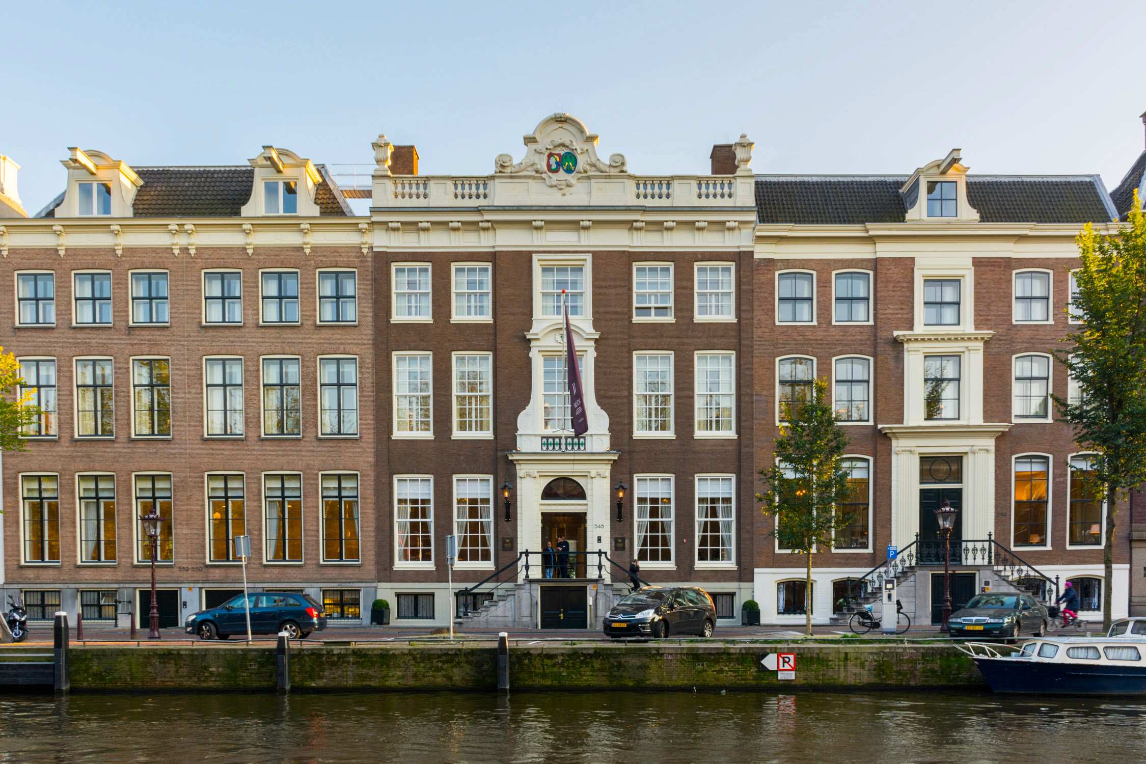 A row of historic brick canal houses with gabled roofs and large windows overlooks a waterway in Amsterdam.
