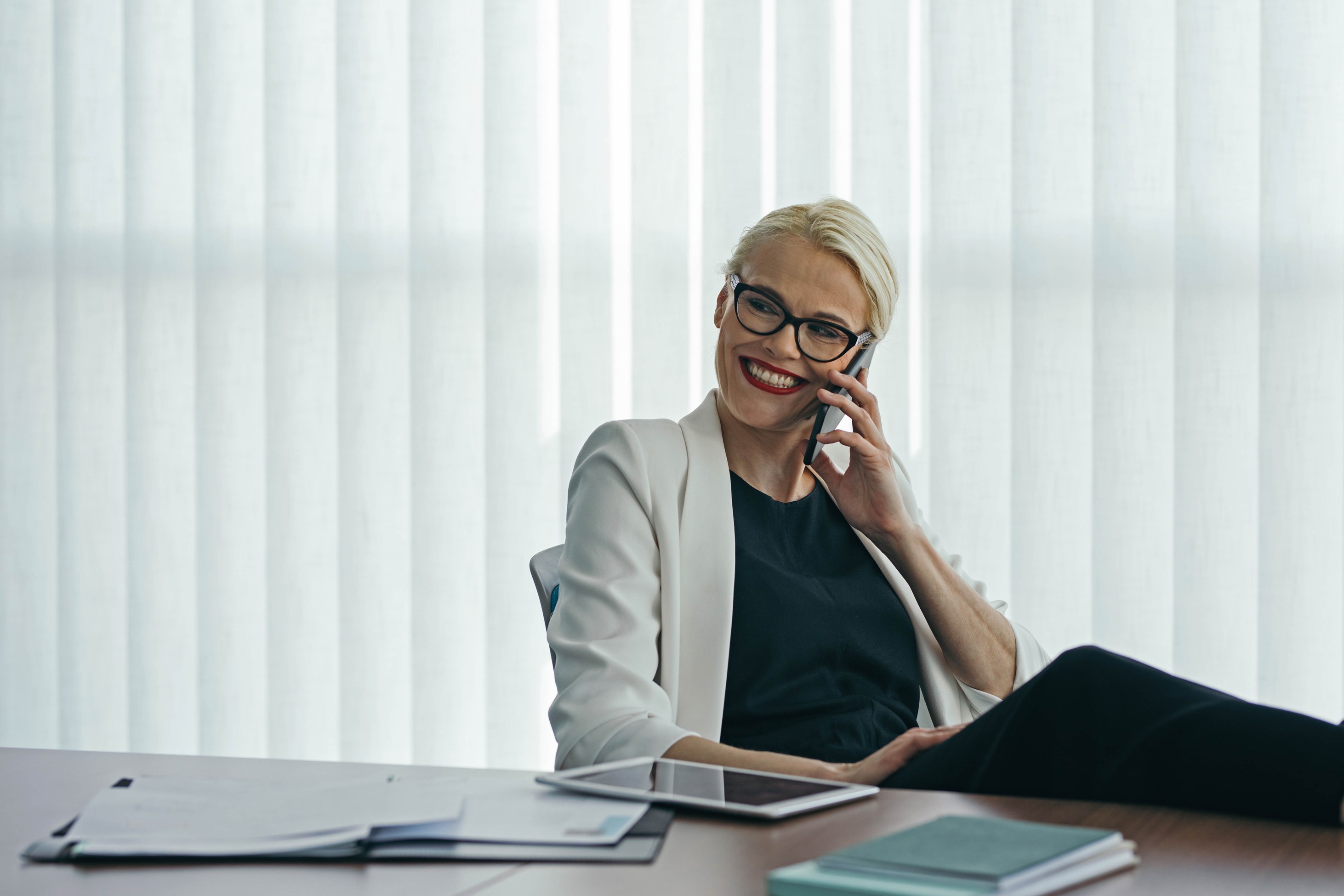 Manager of a recordkeeper talking on the phone, seated at a desk with documents, a tablet, and vertical blinds behind