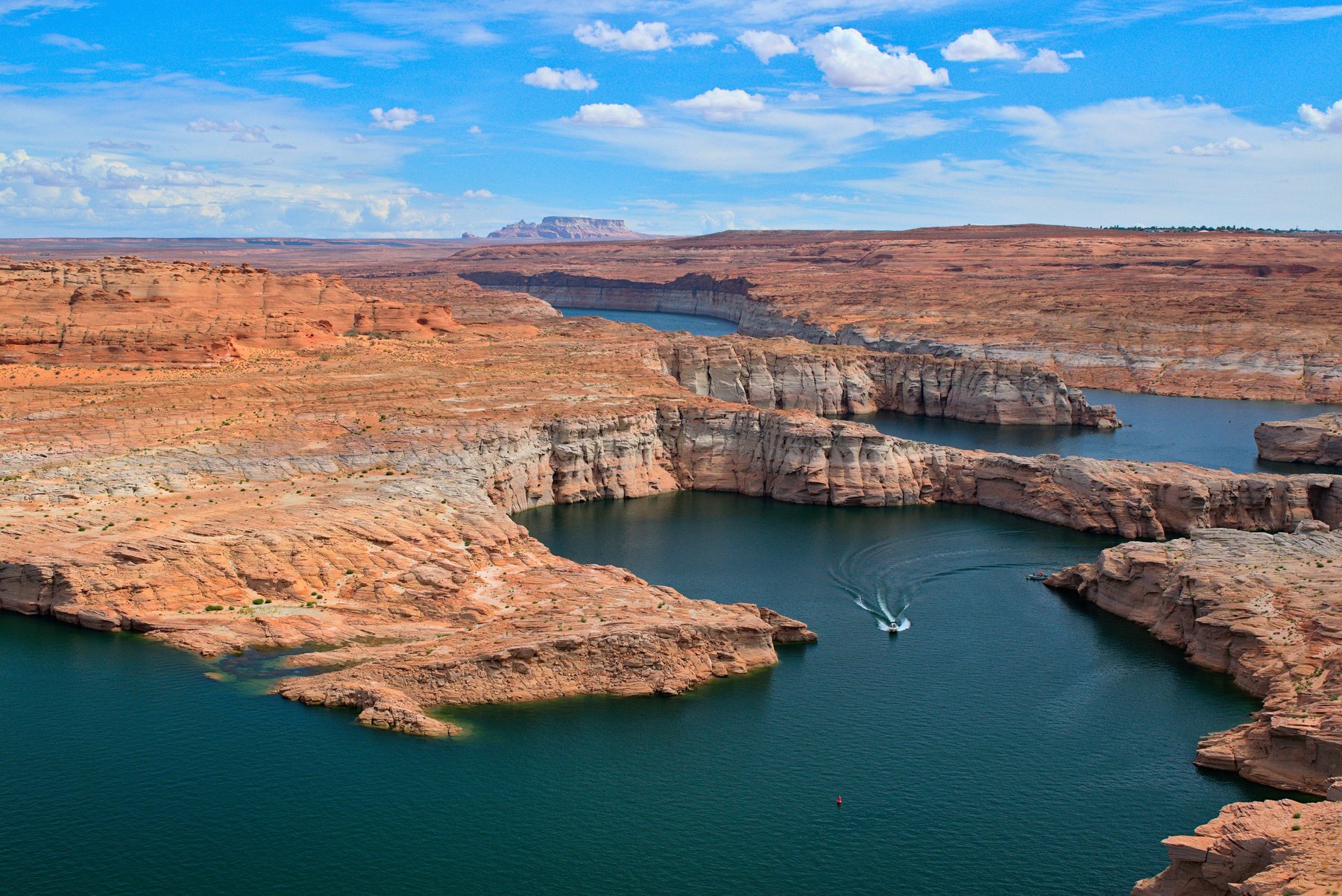 Aerial view of Lake Powell, deep blue water winding through orange and white sandstone canyons under a bright cloud dotted sky, with a single boat leaving a wake.