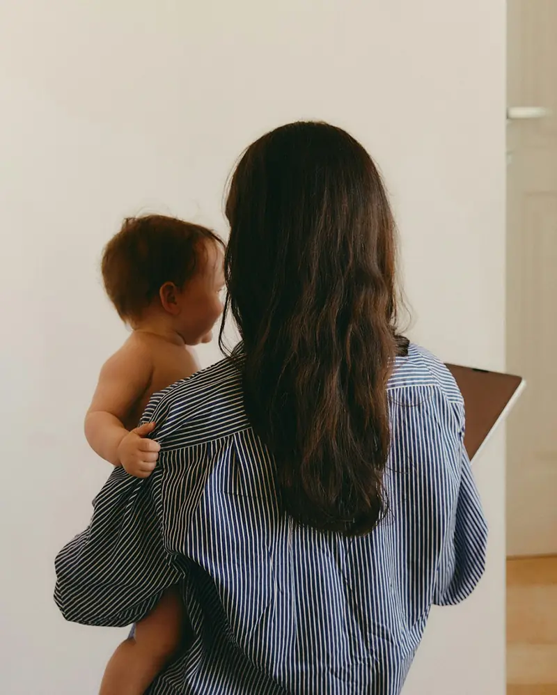Woman seen from behind holding a baby on her shoulder in a softly lit indoor space.