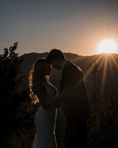 The couple lean their foreheads together in silhouette as sunlight streams over the mountains.