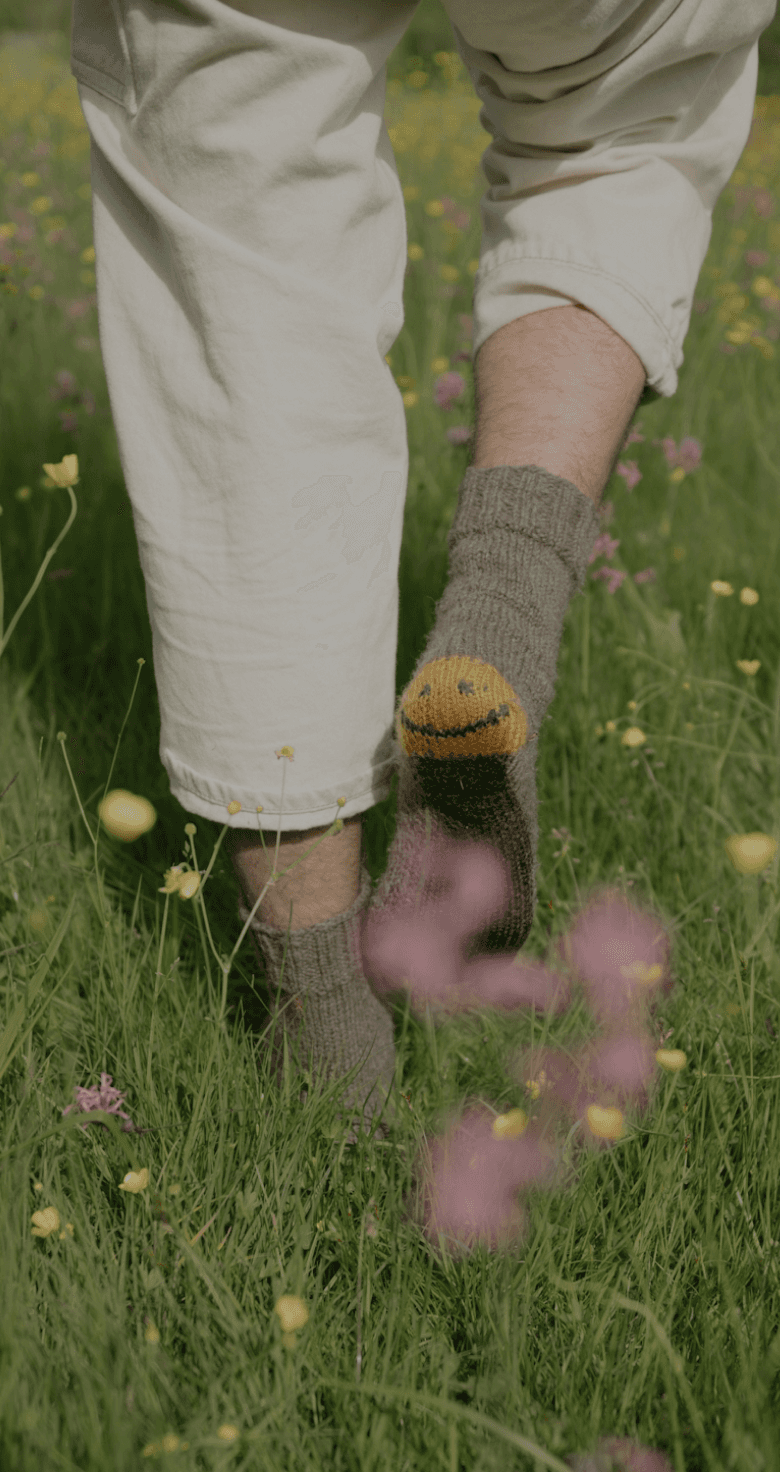 A person's lower body and feet standing in a grassy field filled with yellow and purple flowers, with a knitted sock or leg warmer featuring a smiley face design, creating a serene and whimsical visual composition.