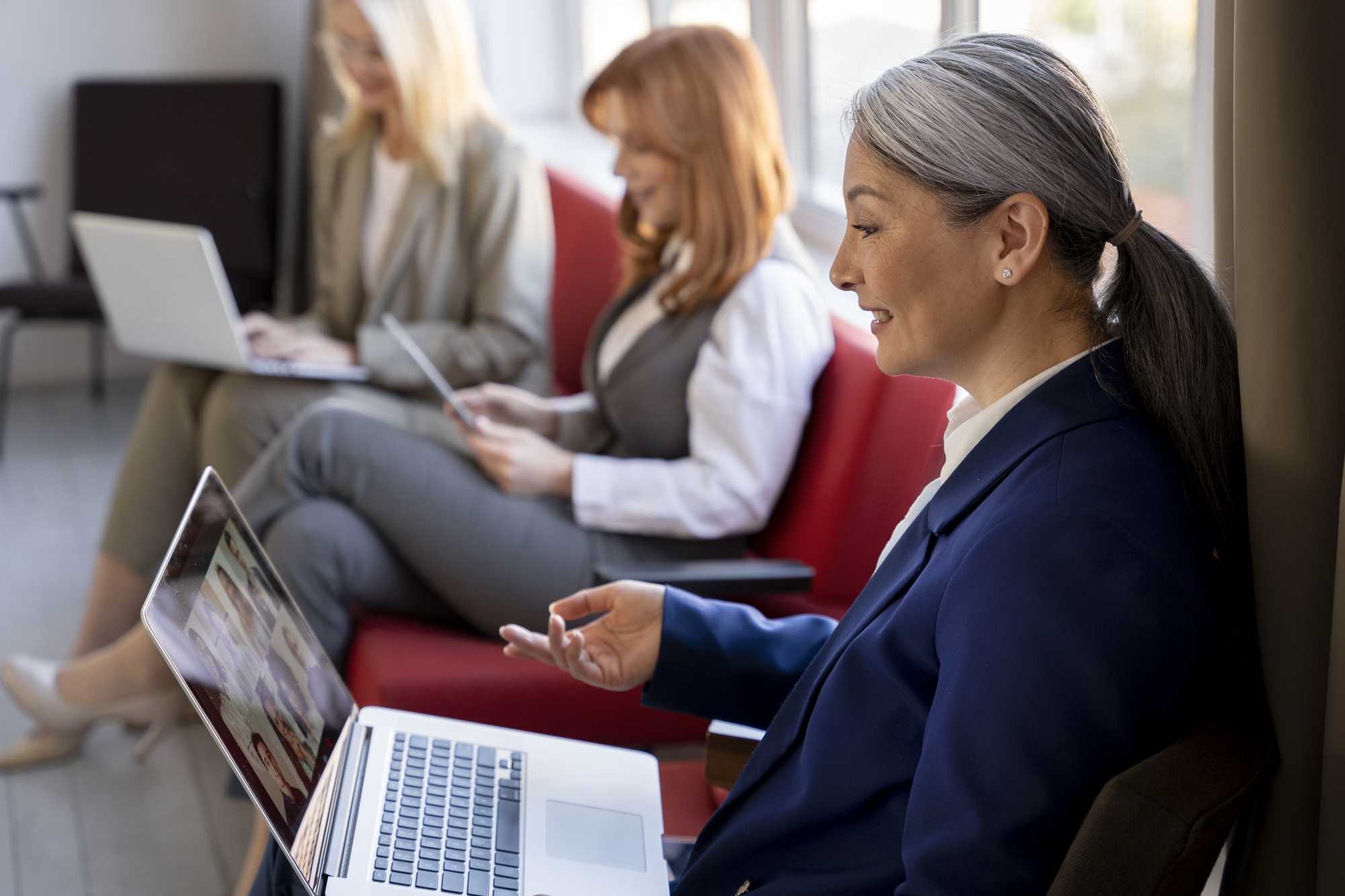 A professional woman with silver hair engaged in a virtual meeting on her laptop, while two colleagues in the background work on a tablet and laptop seated on a red couch