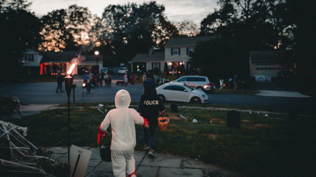 Children trick-or-treating on Halloween at dusk. They wear costumes, one holding a pumpkin bucket. Lit torches line the sidewalk. Houses and trees visible.