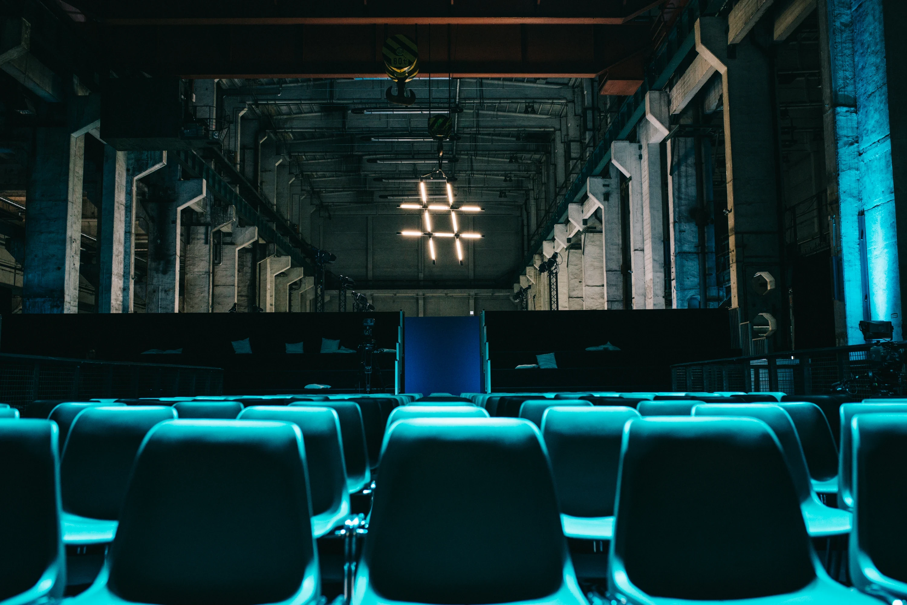 Teal-lit conference seating at FASHIONTECH Berlin facing a glowing hashtag-shaped light sculpture in the long industrial hall.