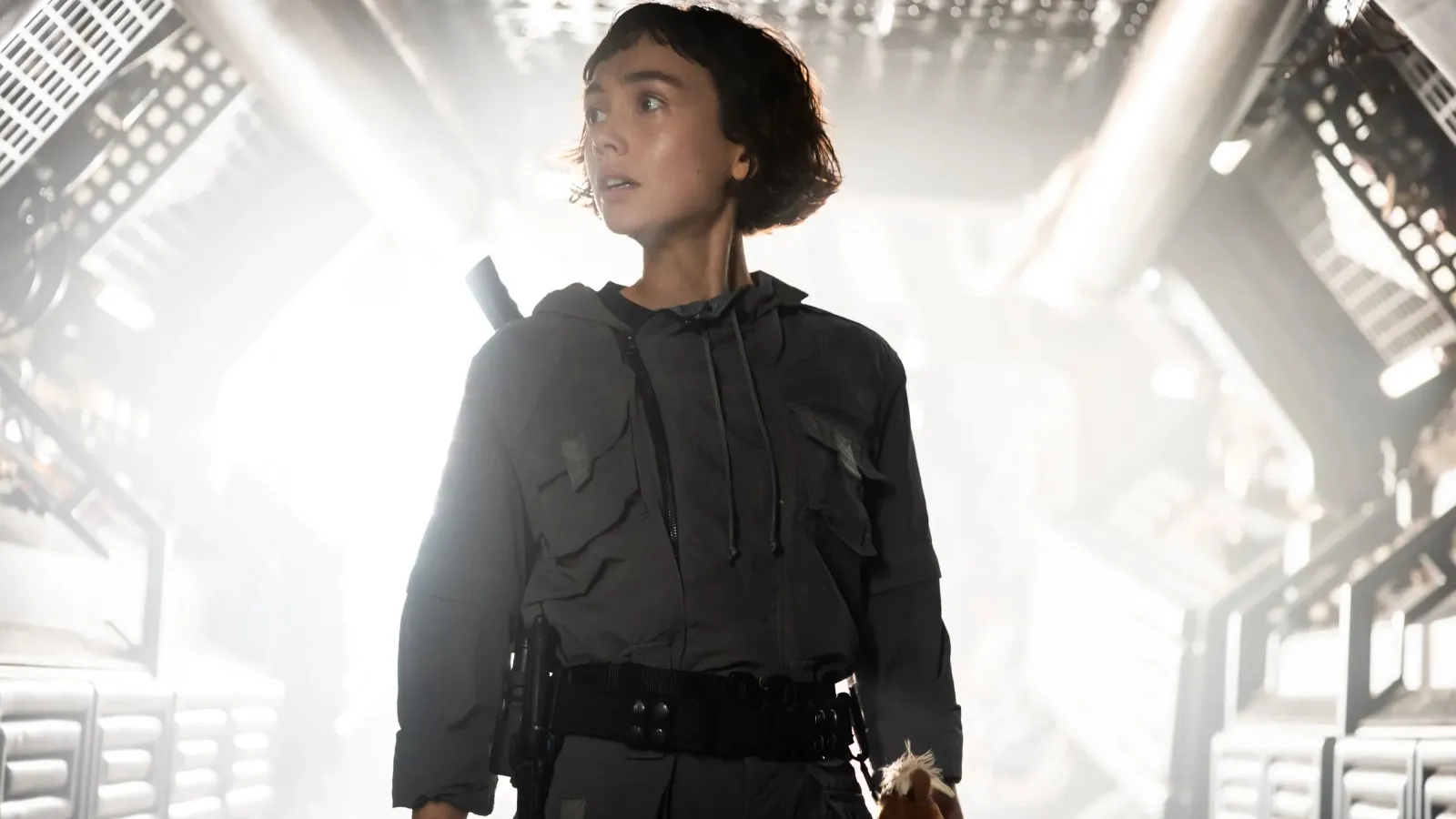 A young woman stands in a dimly lit futuristic corridor wearing a tactical outfit with utility pockets, holding a teddy bear, and exuding a sense of determination and readiness.