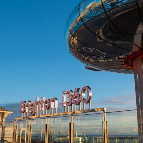 Sign reading "Brighton i360" with the observation tower extending above, set against a blue sky and ocean backdrop.