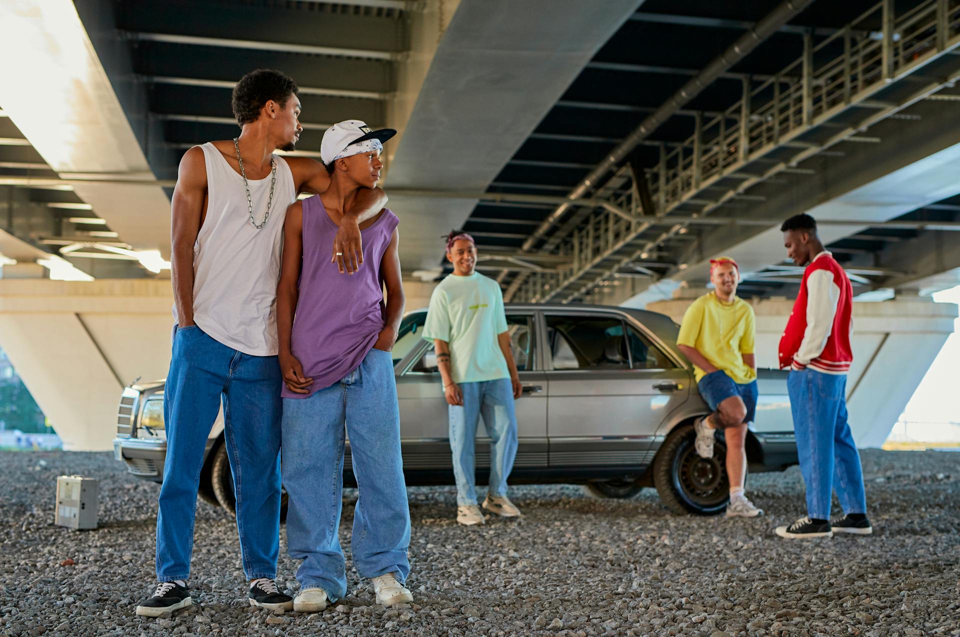 A group of five young men posing outdoors under a bridge with a vintage silver car on a gravel surface.