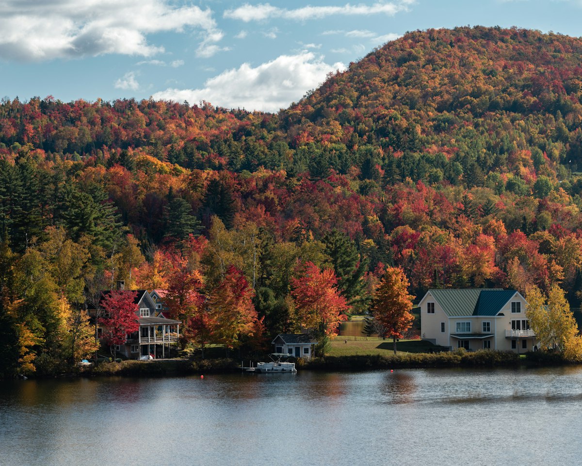Lakeside houses below a hillside of red and orange trees at peak autumn color