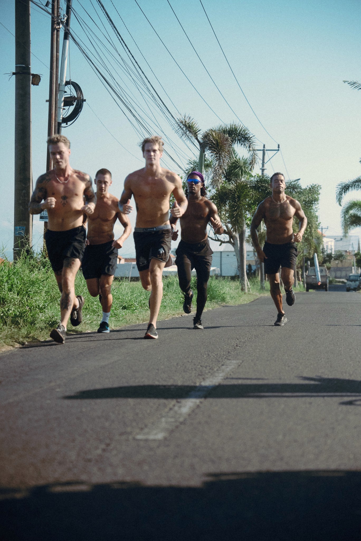 Group of shirtless runners training together on a road in a tropical setting.
