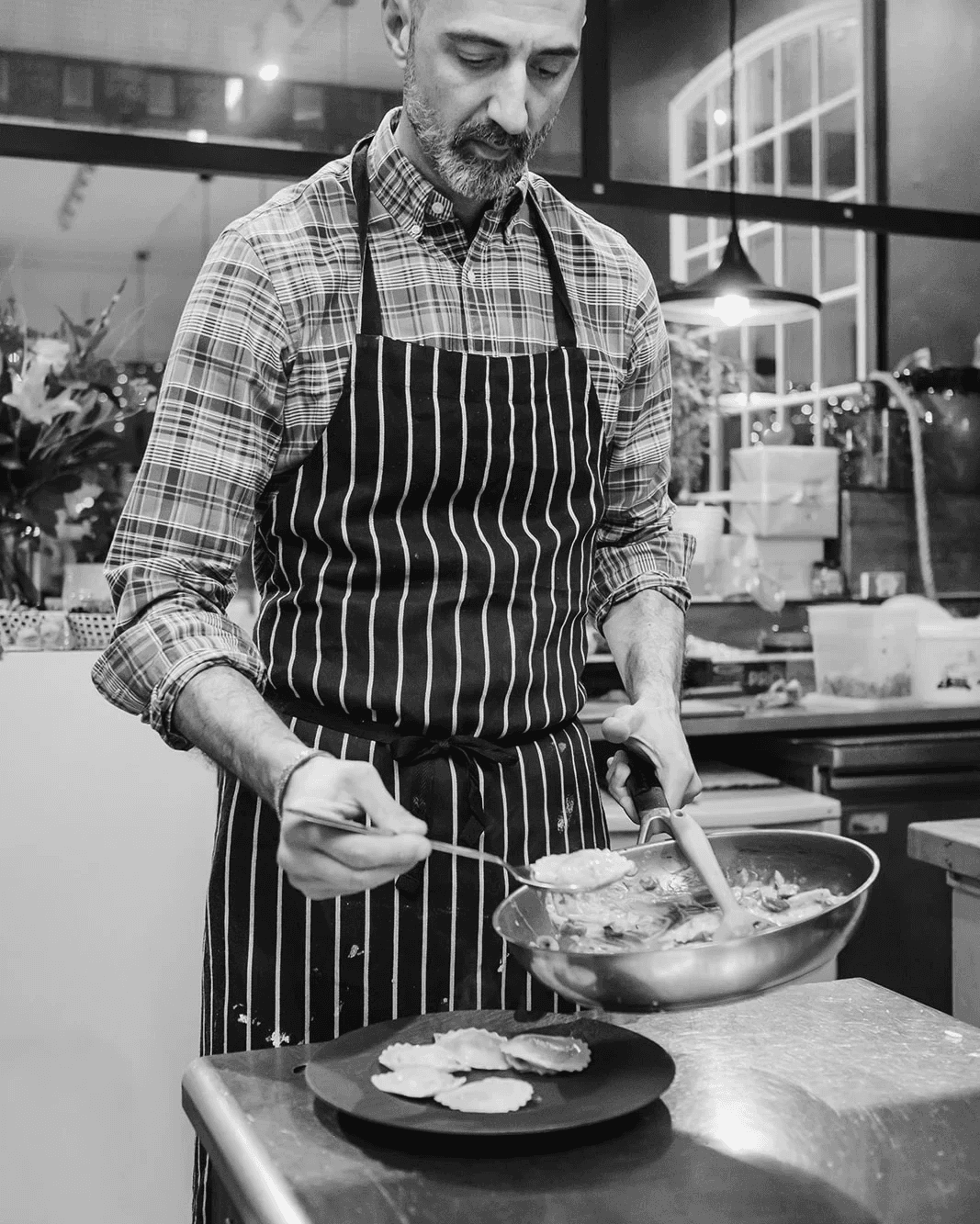 A chef wearing a striped apron and plaid shirt carefully plates fresh ravioli, using a spoon to arrange pieces from a pan in a warmly lit kitchen setting.