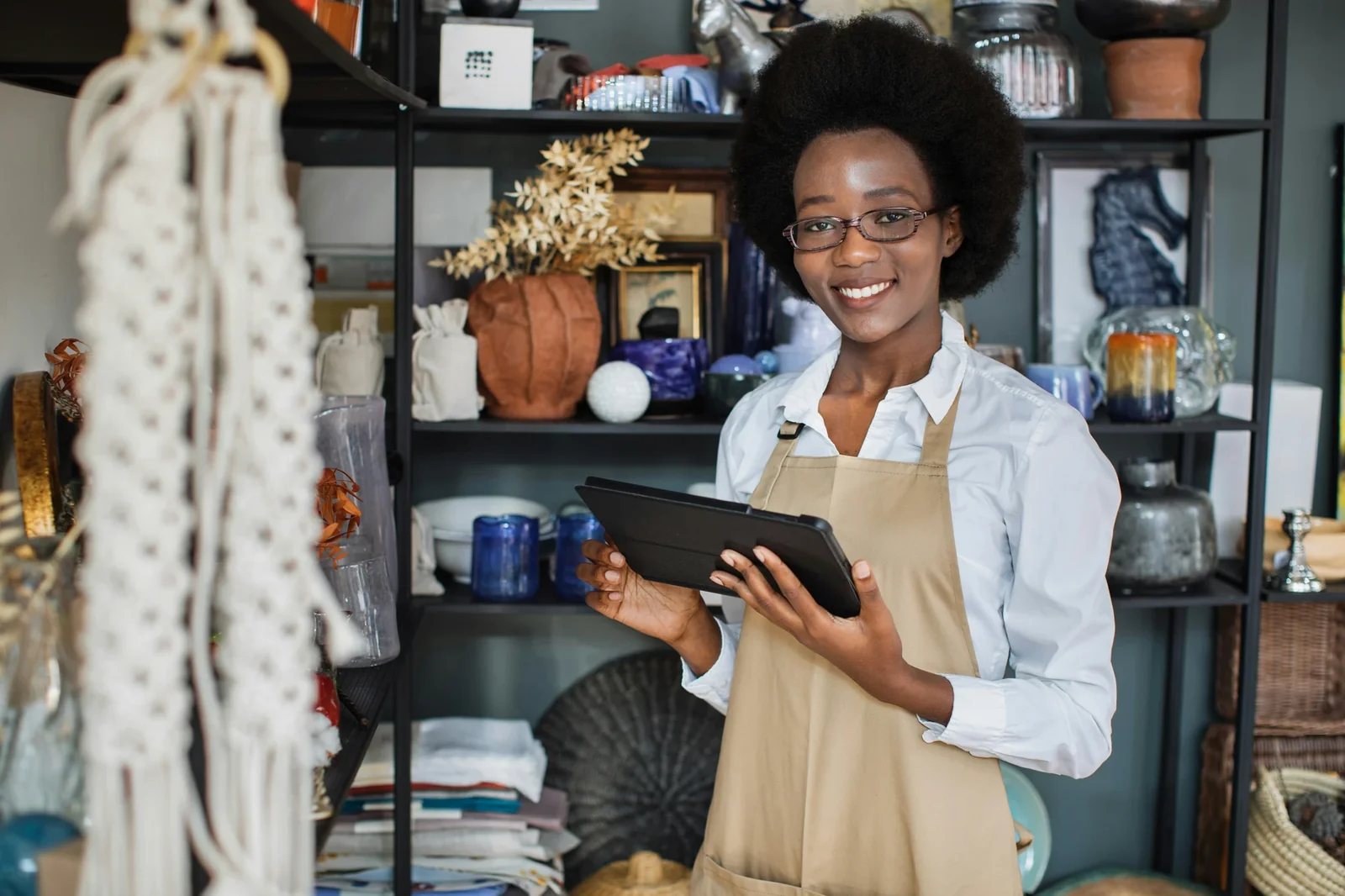 Small business owner wearing apron holds tablet while smiling in front of shelving displaying home décor products.
