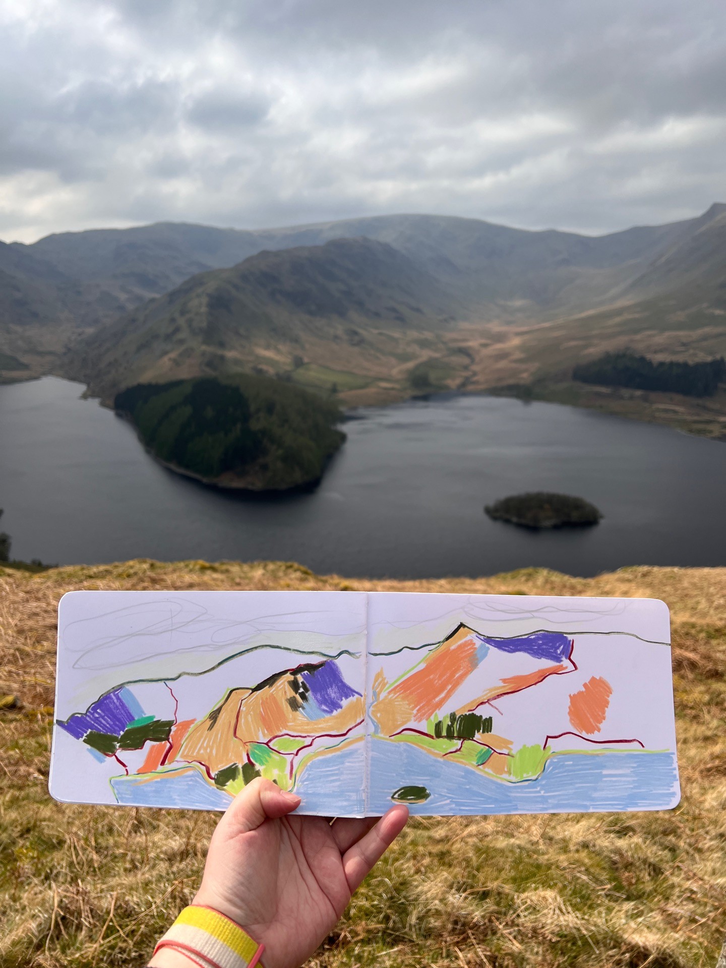 An illustration in April's sketchbook that looks over the reservoir. The water in the foreground is surrounded by the fells around, with an outcrop filled with trees.