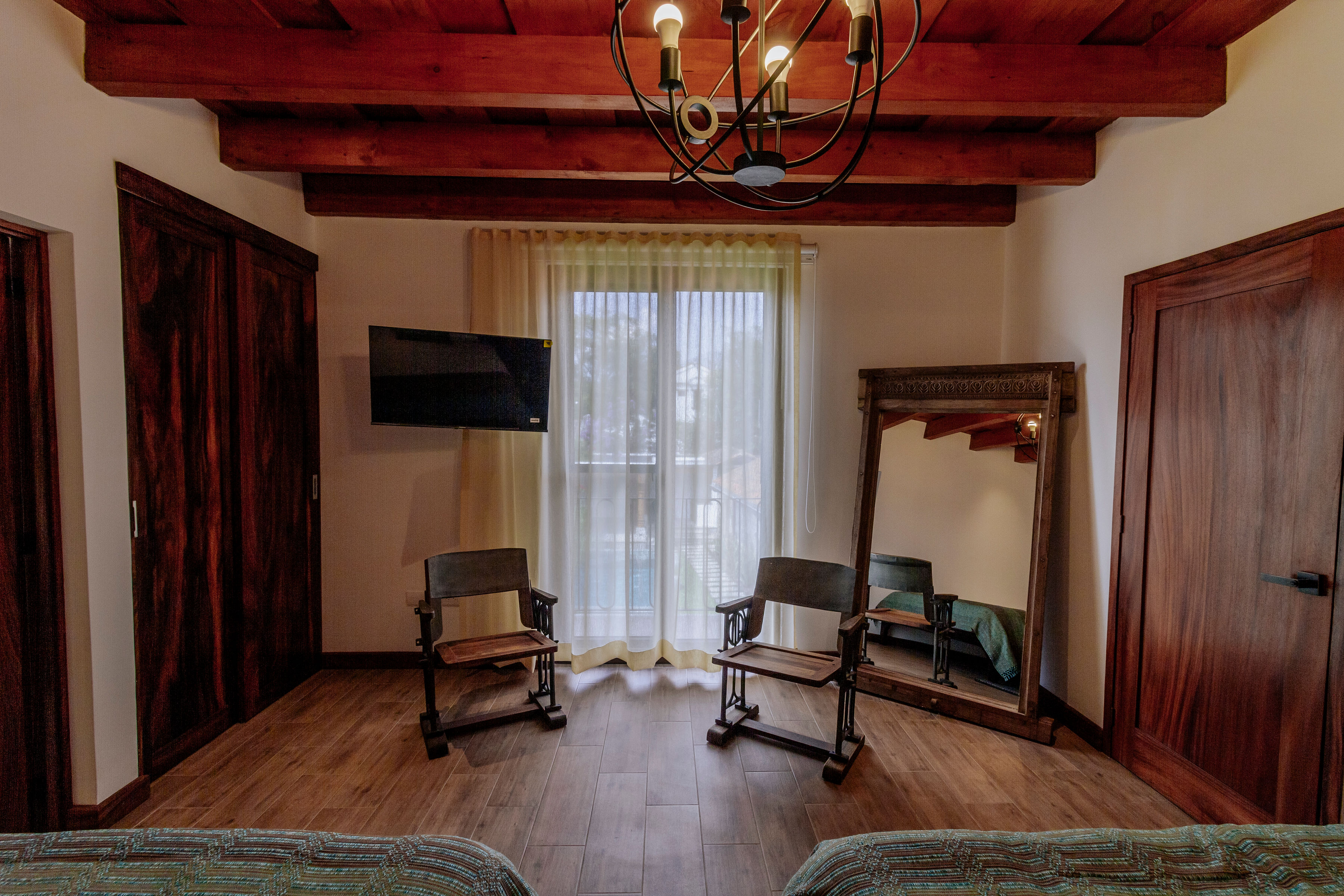 A cozy, rustic bedroom features wooden beams on the ceiling, two wooden chairs, a large mirror, and a wall-mounted TV, with natural light streaming in through sheer curtains.
