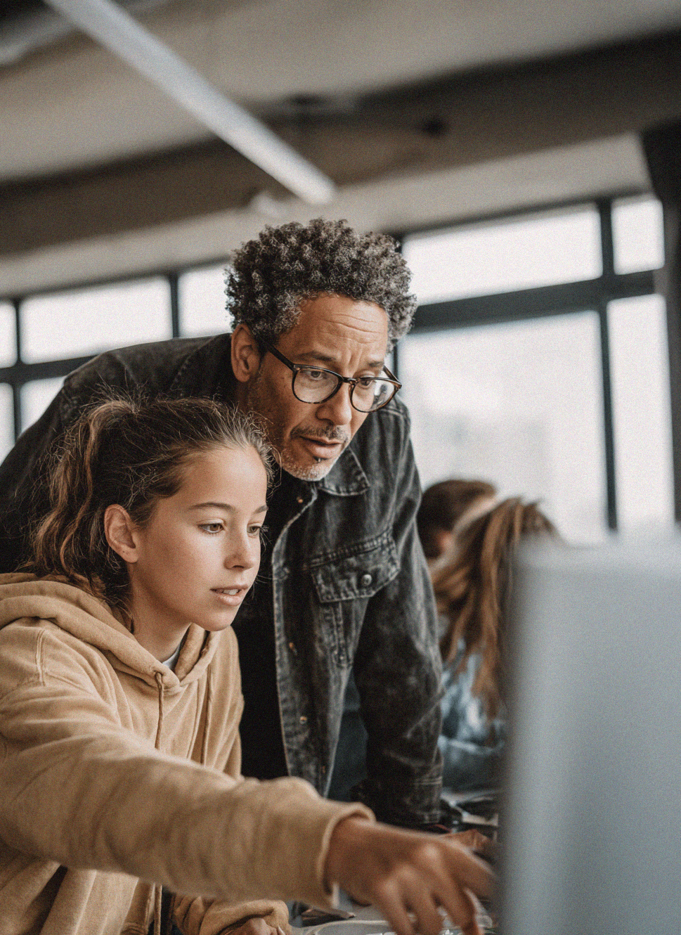 A young person wearing a tan hoodie sits at a desktop computer, focused on the screen while pointing toward something displayed. An older adult wearing glasses and a dark jacket leans in beside them, looking at the monitor and appearing to provide guidance. The setting appears to be a modern classroom or workspace, with another person working at a computer in the background.