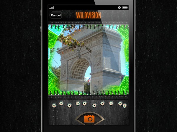 The image shows a smartphone screen displaying a photo editing application. The application interface includes a camera icon in the bottom center and a grid of camera controls below it, including circular buttons and a flash icon. The photo displayed on the phone’s screen is a white archway, possibly the Arc de Triomphe, with green foliage visible around it. The background of the phone screen is black with a green border around the image. The screen also displays “WILDVISION” at the top center and a “Cancel” button in the top left corner.