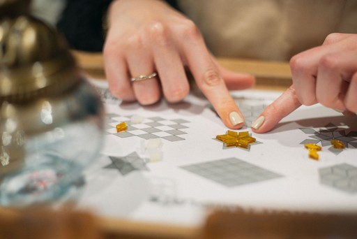 A close-up of hands organizing mosaic tiles on a design template before placing them on a lamp, emphasizing the planning and precision involved in mosaic art.