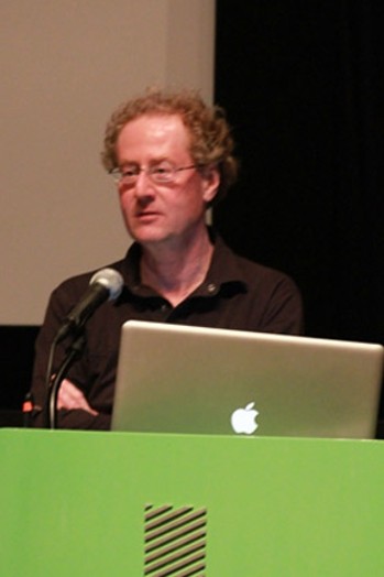 A man with glasses giving a lecture at a green stand.