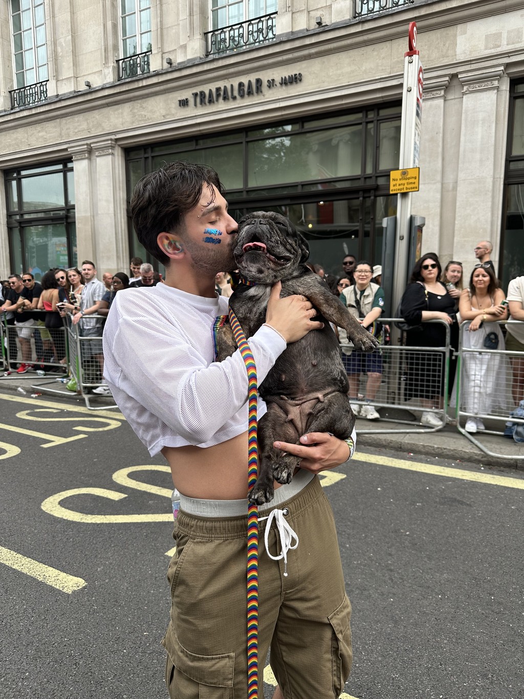 A person standing on a city street lovingly holds and kisses a joyful dog, capturing a playful moment outside a building named "The Trafalgar St. James," surrounded by a crowd, embodying the spirit of dog adventures.