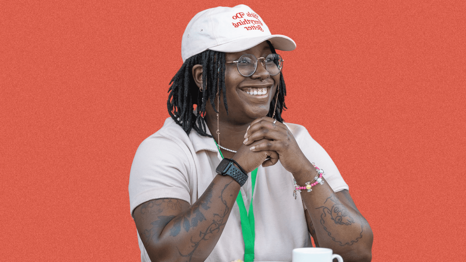 Smiling person with curly hair and glasses wearing a white cap and shirt, against an orange background.