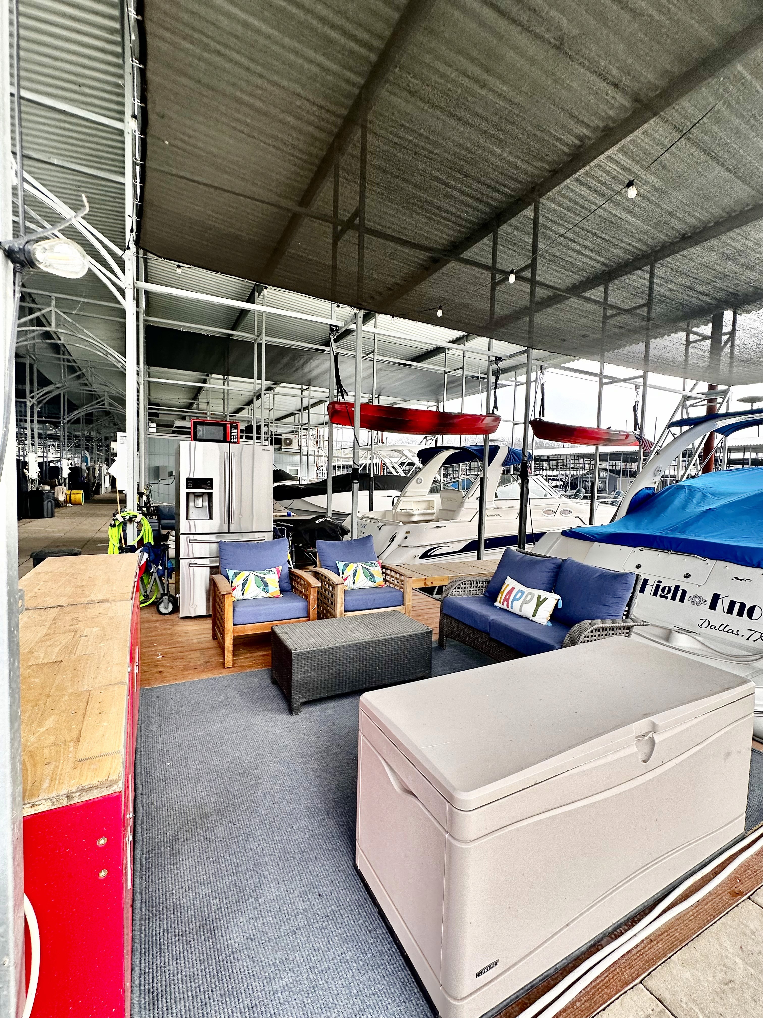 A cozy marina lounge features comfortable seating with colorful cushions, adjacent to multiple moored boats beneath a sheltered, industrial-style roof.