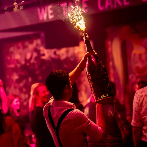 Seorang pengunjung memegang botol besar dengan kembang api di klub malam yang ramai dan remang-remang dengan pencahayaan merah muda dan merah.
