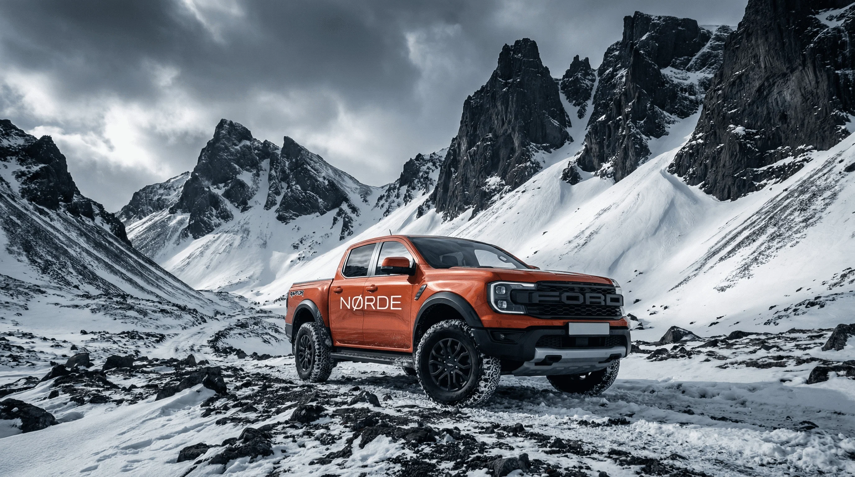 An orange Ford Ranger Raptor pickup truck parked on a snowy, rocky landscape with dramatic mountains in the background.