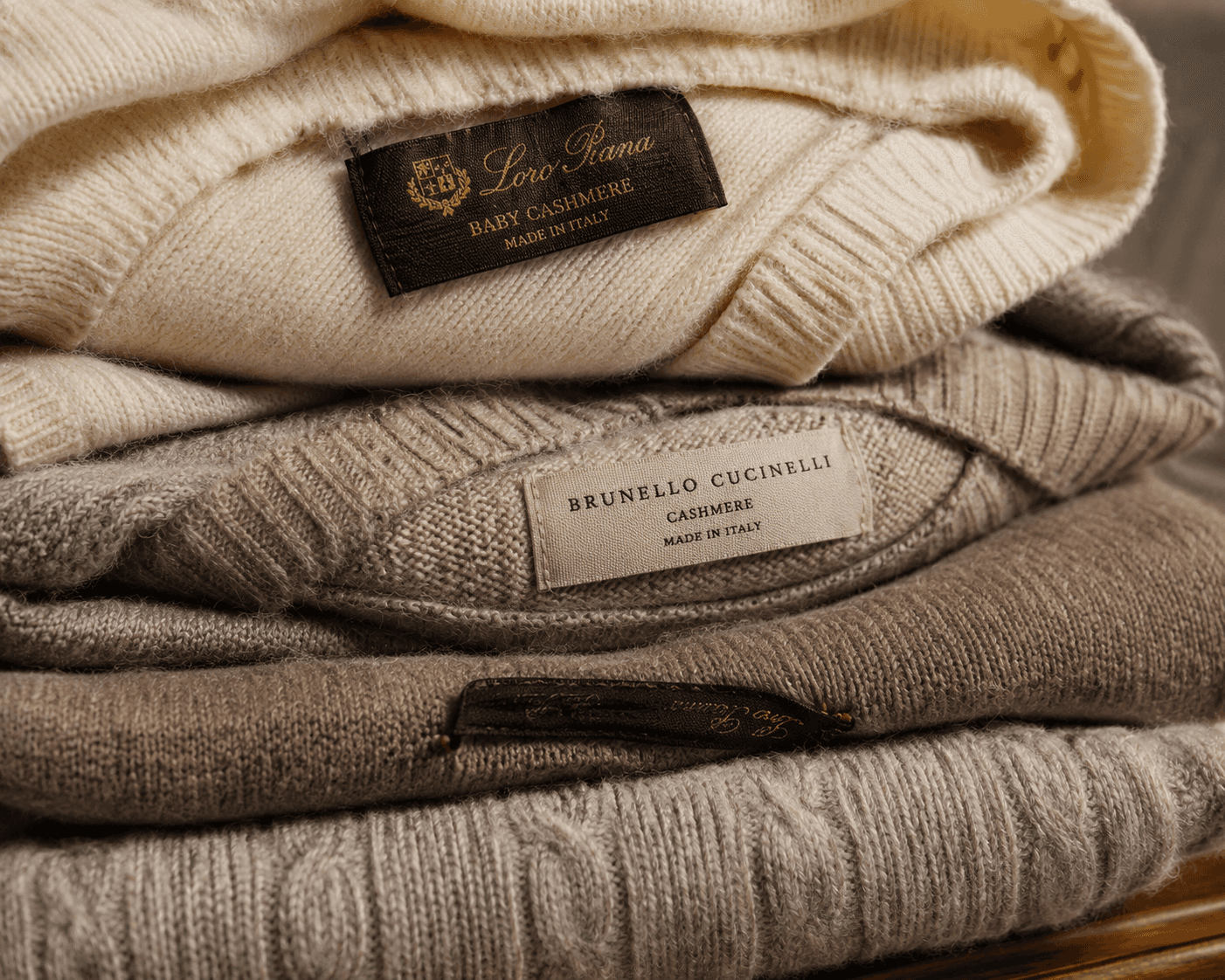 Close-up stack of luxury Italian menswear knits, featuring a cream-colored Loro Piana baby cashmere sweater on top, layered over a beige Brunello Cucinelli textured knit cashmere sweater, followed by neutral brown and gray cable-knit wool layers showing their brand labels.
