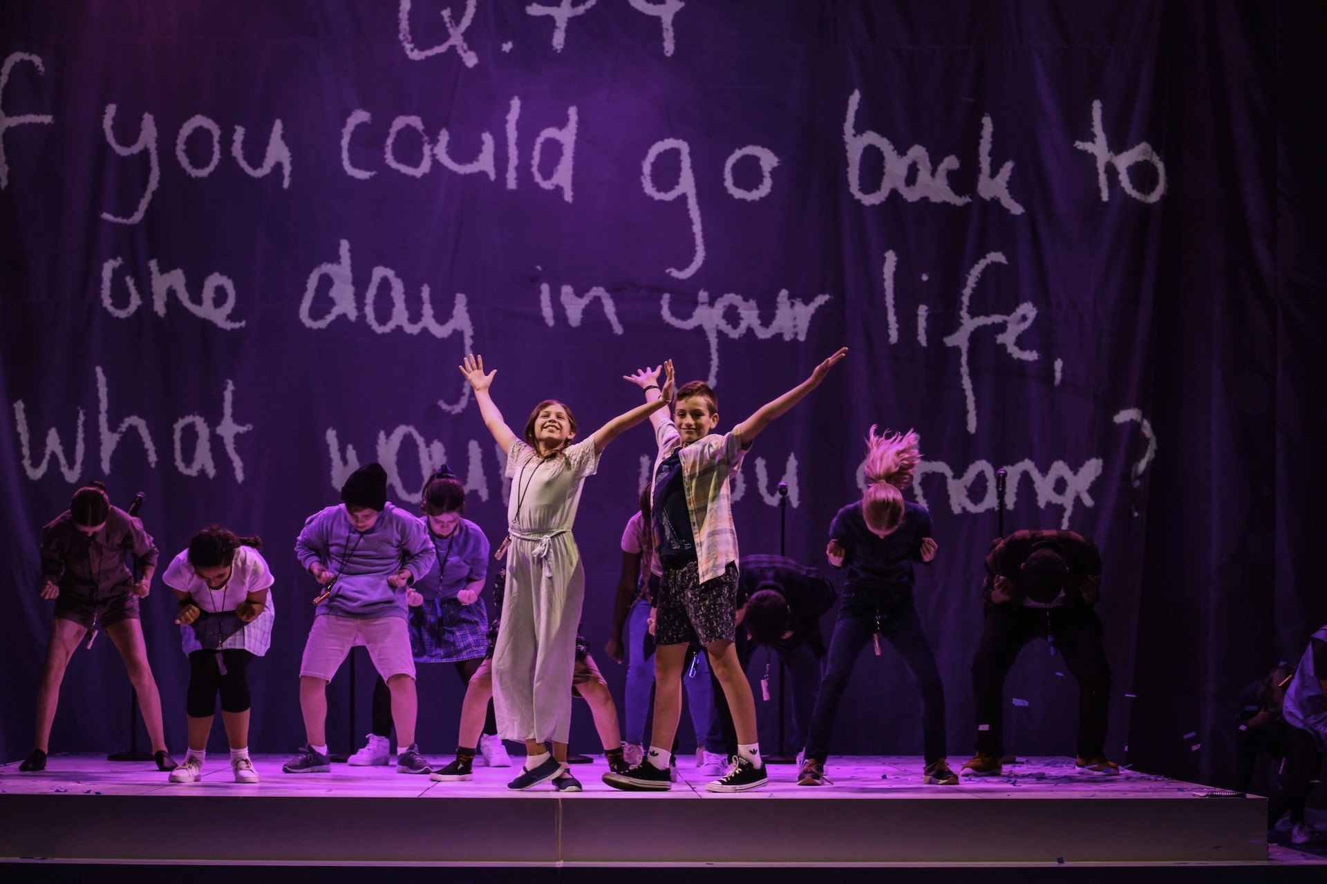 Two young performers stand centre stage with arms stretched wide in a joyful pose, while the rest of the cast bends forward behind them. A large projected question fills the backdrop, reading, “If you could go back to one day in your life, what would you change?”, creating a playful and reflective moment in the show.