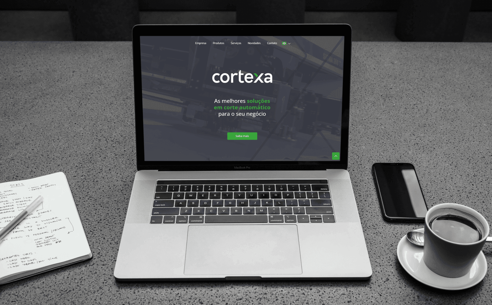 Notebook com o site da Cortexa aberto, exibindo o texto 'As melhores soluções em corte automático para o seu negócio', ao lado de um caderno com anotações, um smartphone e uma xícara de café sobre uma mesa de concreto.