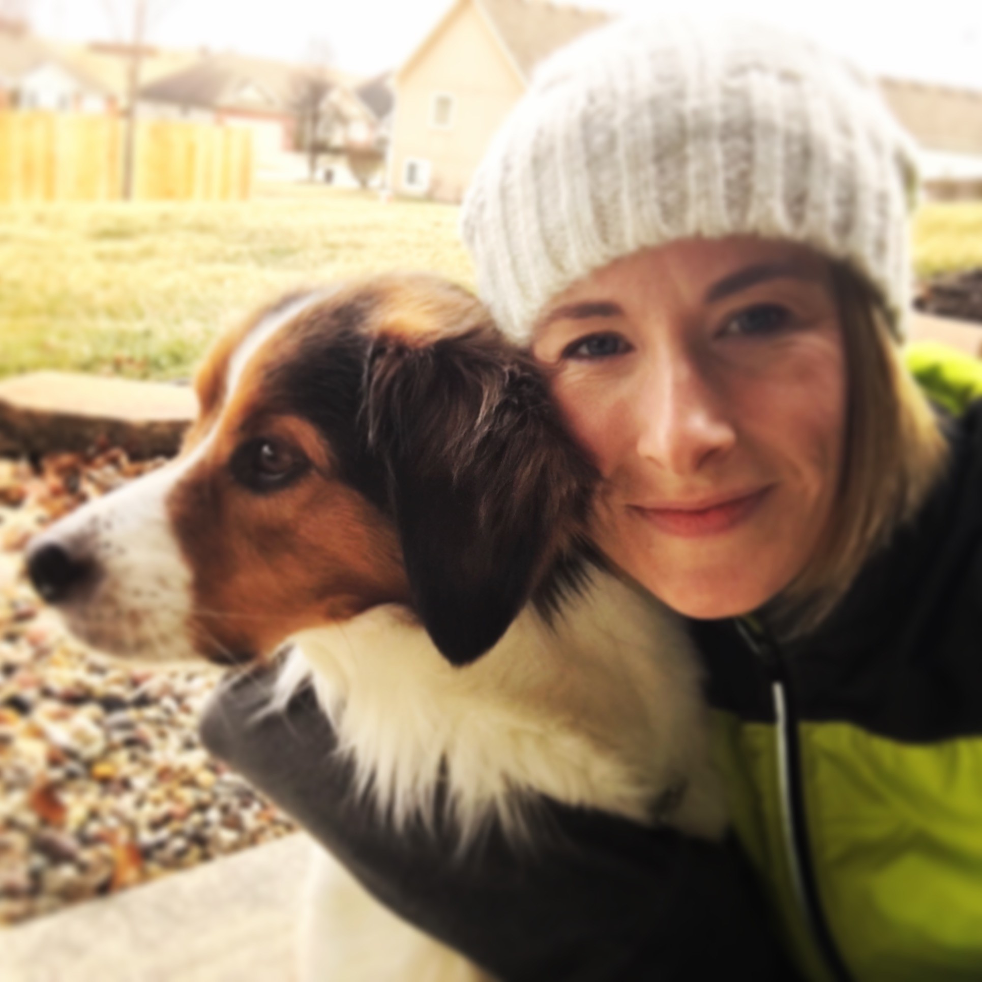 Heather Hausman wearing a knit hat and green jacket snuggles with a brown and white dog outside on a grassy yard with houses in the background.