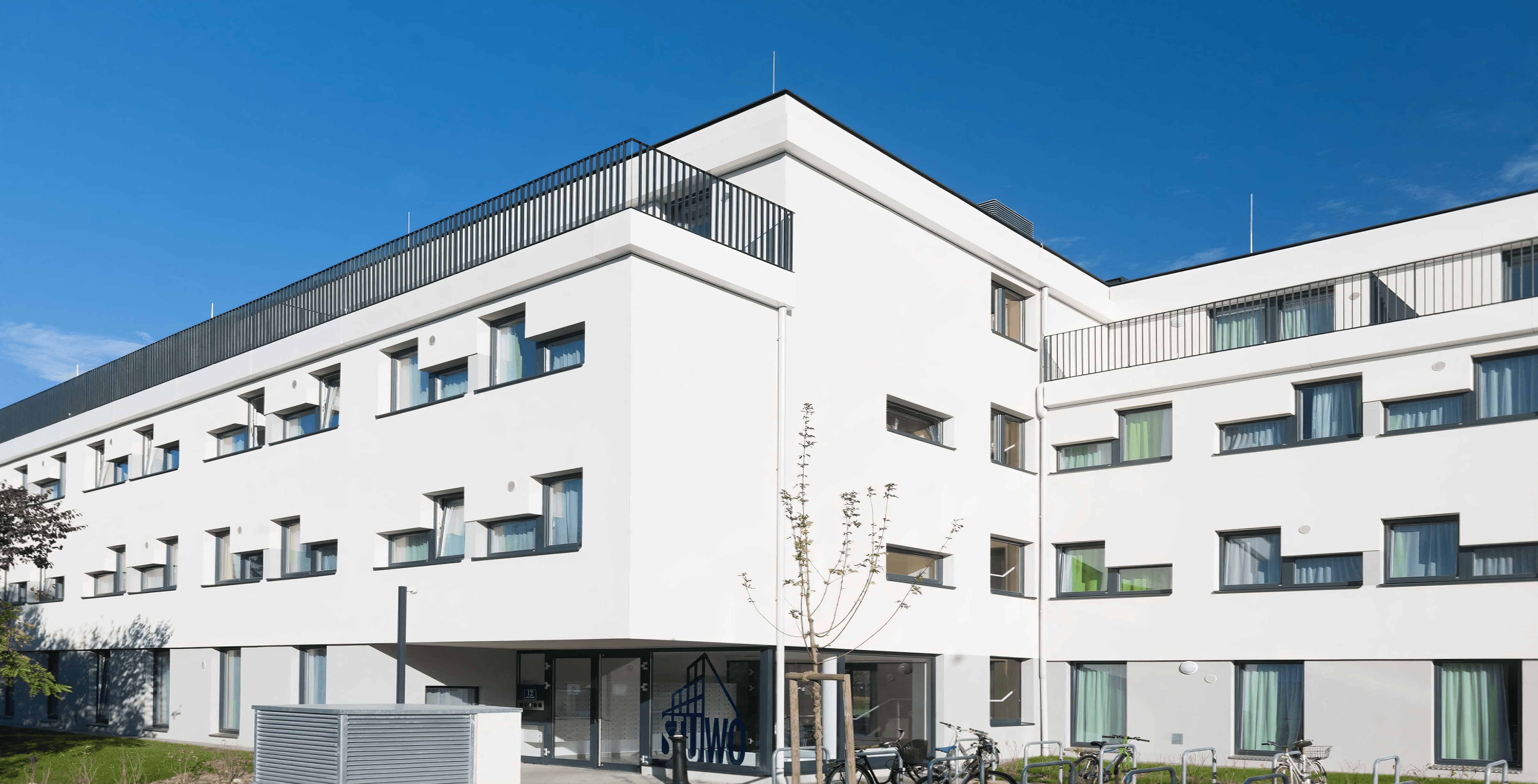 MW Interior - Modernes weißes STUWO Studentenwohnheim mit mehreren Stockwerken, Dachterrasse, grünen Vorhängen hinter Fenstern und Fahrradabstellplätzen" Beschreibung: Klares, zeitgemäßes Studentenwohnheim der STUWO mit weißer Putzfassade, Dachterrassengeschoss und begrüntem Eingangsbereich – ein Referenzstandort, für den sämtliche Zimmer- und Gemeinschaftsflächeneinrichtungen realisiert wurden.