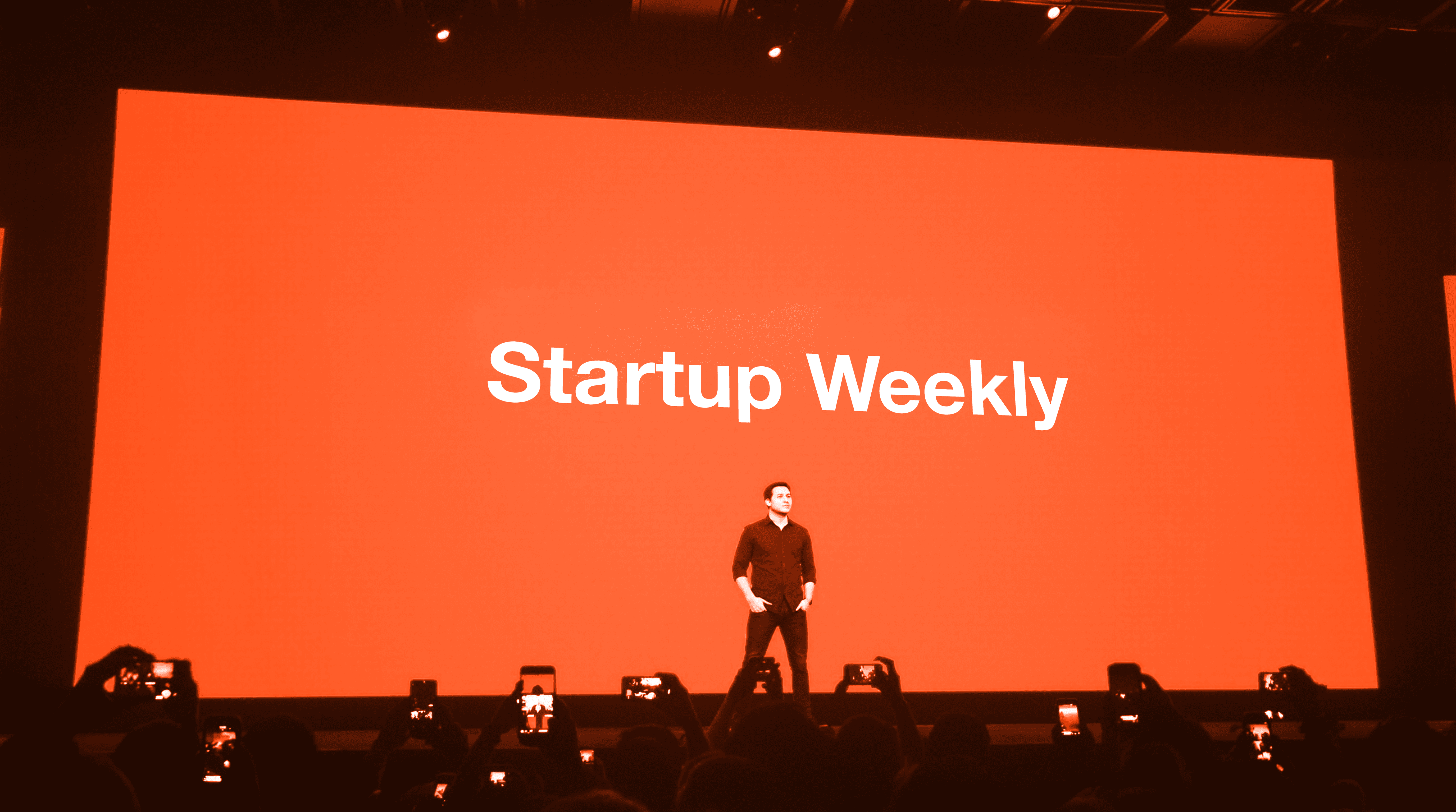 A speaker stands on stage in front of a bright orange screen displaying the text "Startup Weekly."