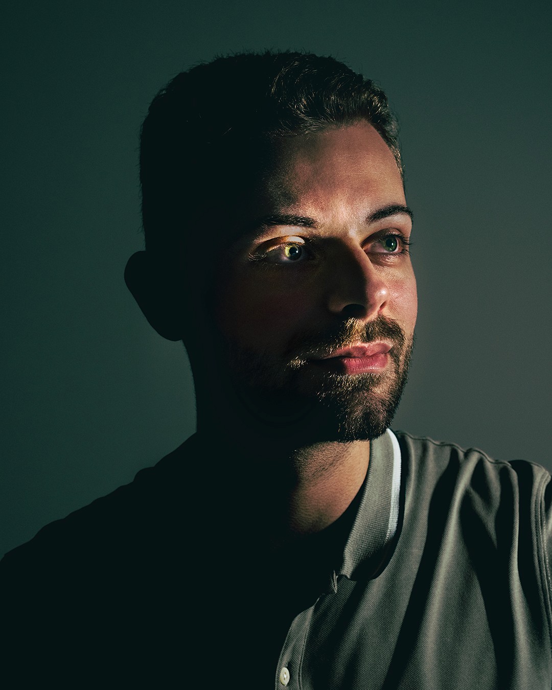 Portrait d’un homme éclairé en clair-obscur, le visage partiellement illuminé par une lumière latérale qui met en valeur ses yeux et son expression pensive. L’arrière-plan est sombre, créant un contraste fort.