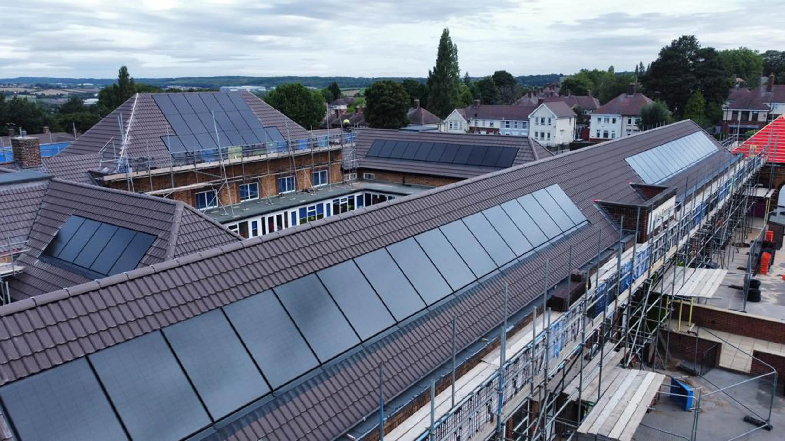 Aerial view of a large building complex under construction, featuring extensive rooftop solar panels on sloped tile roofs, surrounded by scaffolding and set against a backdrop of suburban houses and greenery under a cloudy sky.
