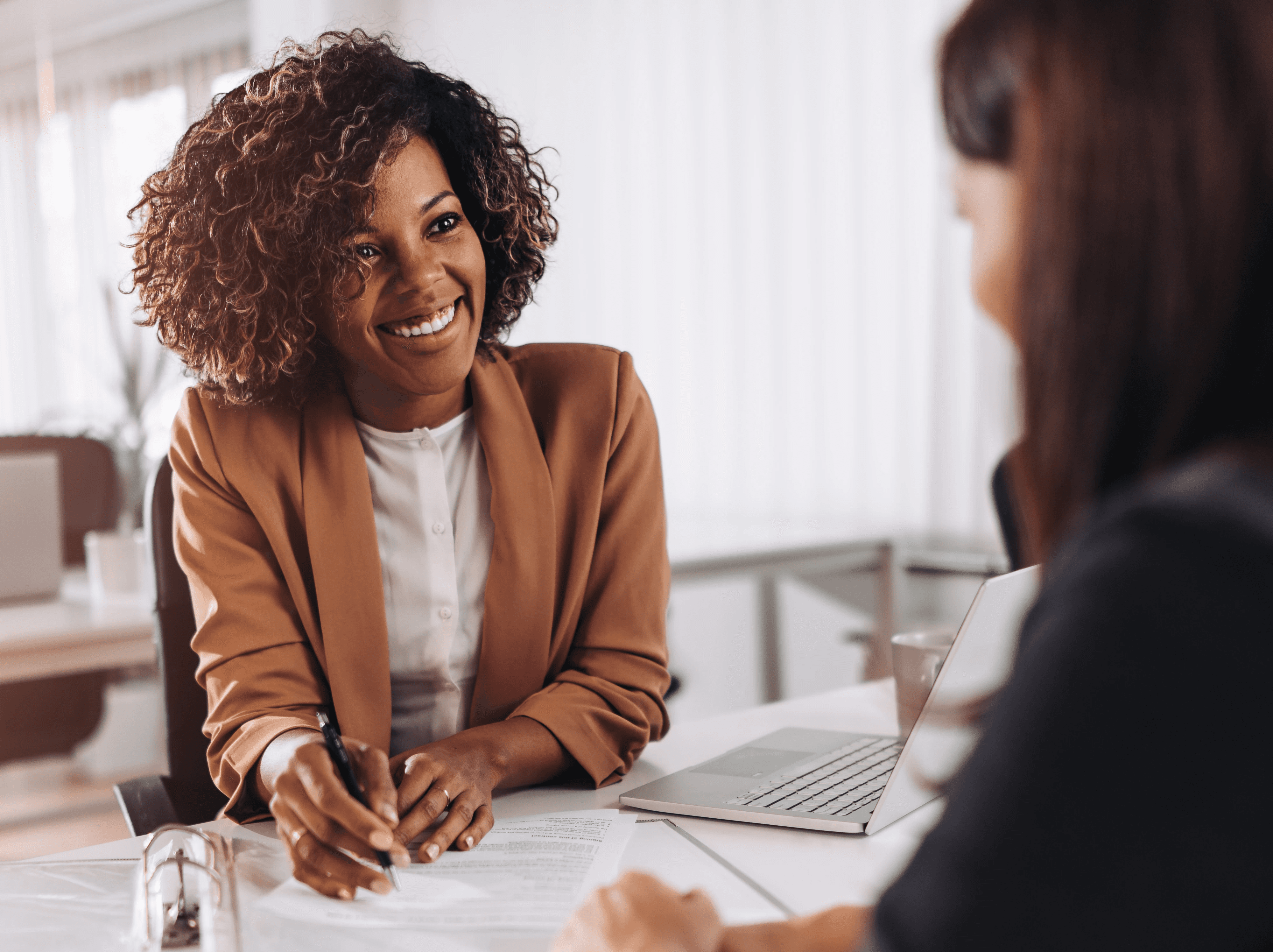 Lächelnde Geschäftsfrau im braunen Blazer sitzt an einem Schreibtisch und führt ein Gespräch mit einer Kollegin in einem hellen Büro.