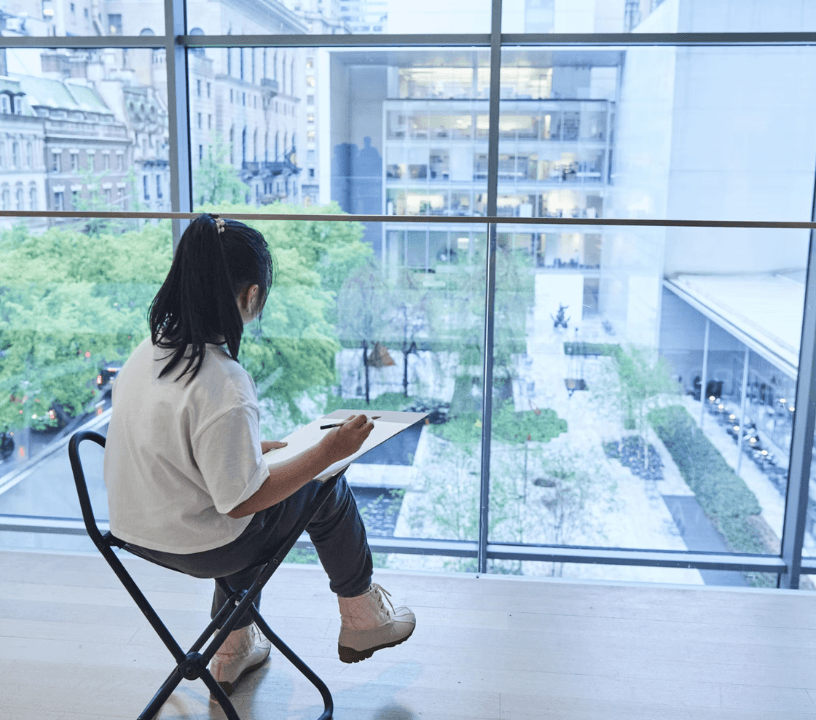 An artist overlooks the courtyard of MoMA sketching. The trees are green and the sun us shining.