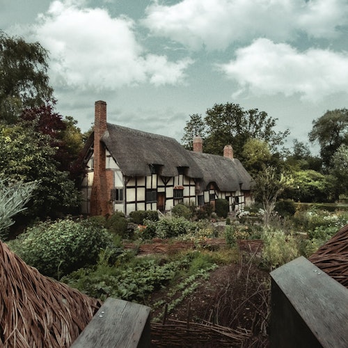 Olkipäällysteinen mökki, jota ympäröi vehreä kasvusto ja puutarhat osittain pilvisen taivaan alla.