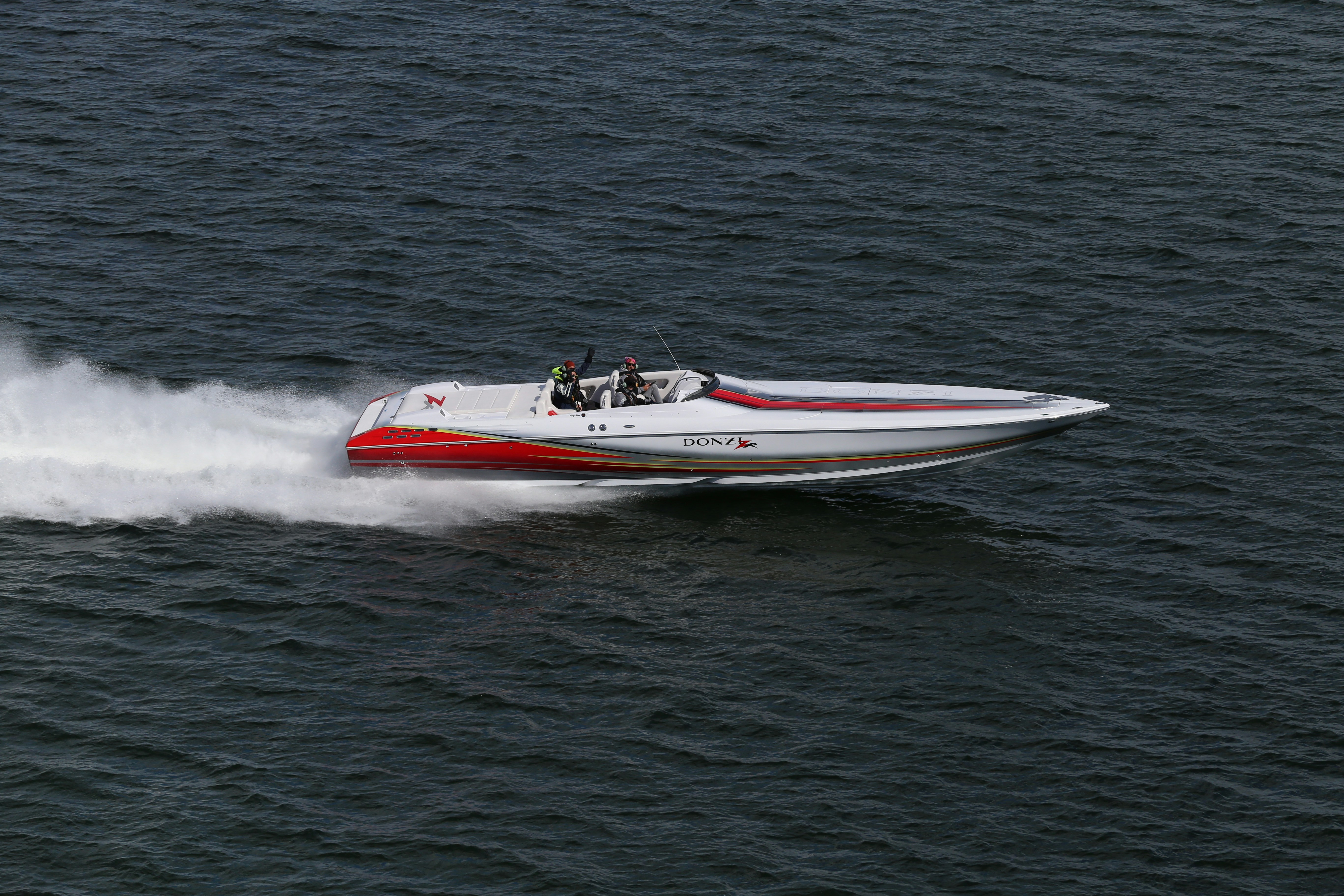 A sleek, high-speed powerboat with a red and white design races across the open water, leaving a trail of white spray in its wake.