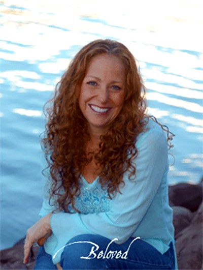 A smiling woman with her arms crossed, standing against a dark green background. She has long, dark hair.