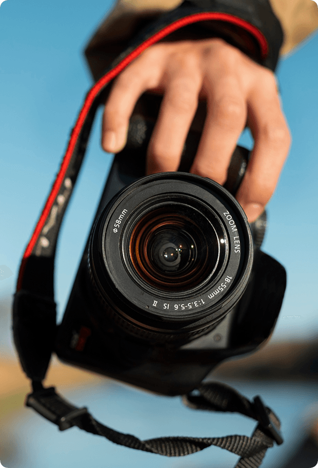 A person with curly hair holds a vintage camera to their eye, set against a dark background. Blue and pink lighting creates a dramatic, focused mood.