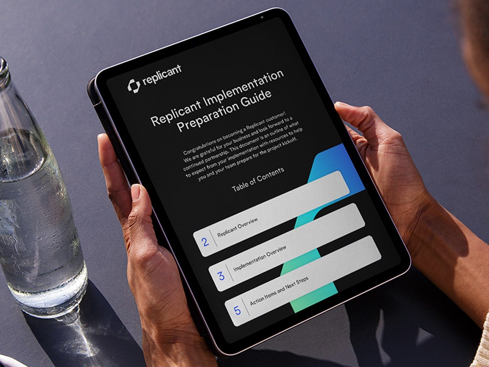 Person holding a tablet displaying the Replicant Implementation Preparation Guide, featuring branded onboarding documentation with structured layouts, bold typography, and modular interface elements in Replicant’s visual identity system.