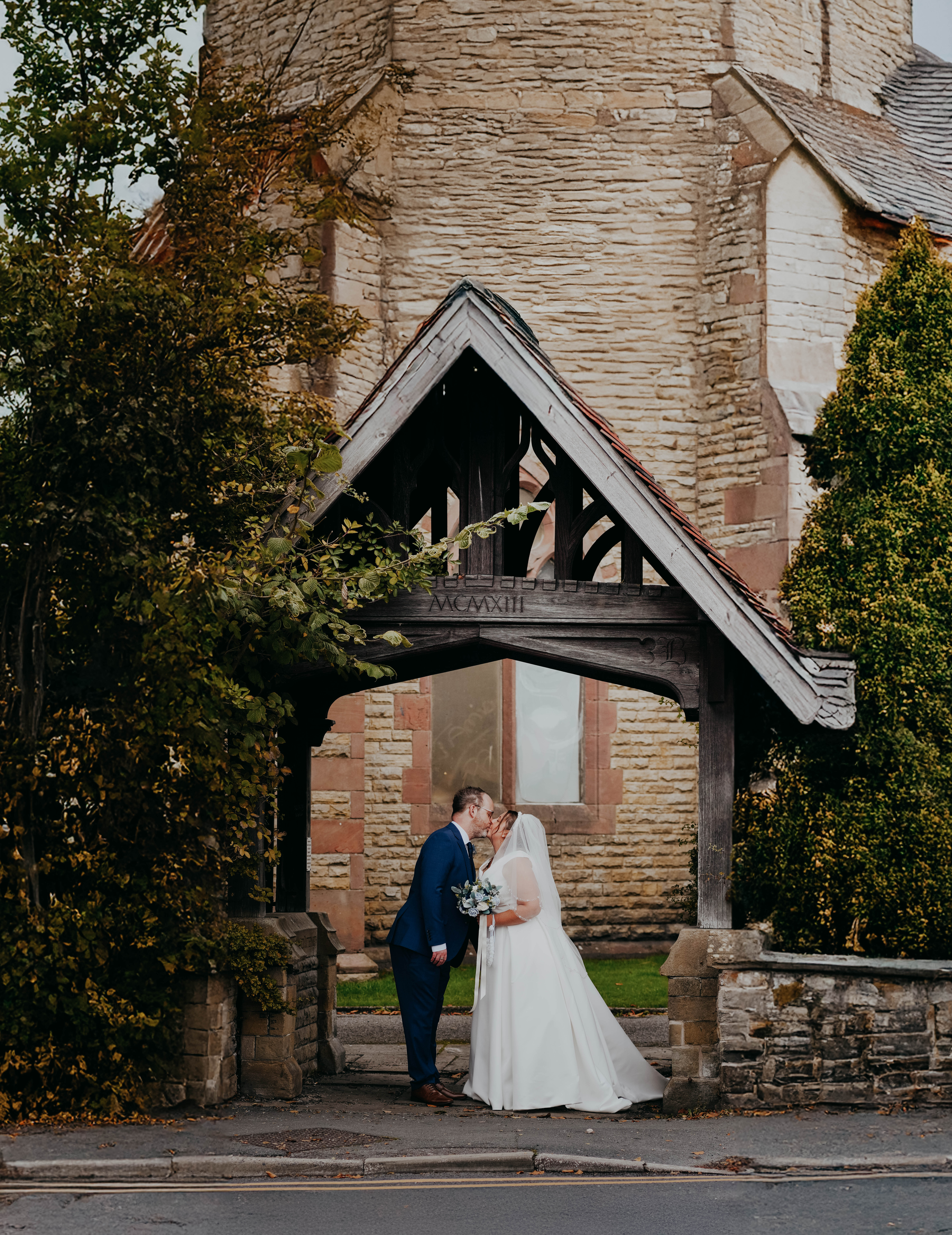 The bride and groom share a kiss while standing under the wooden archway entrance to a stone church. The bride is in her white gown and veil, and the groom is in his blue suit.