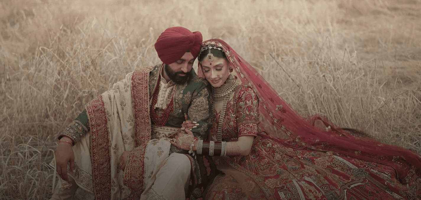 Man and woman sitting in a field, wearing burgundy wedding attire with light brown and gold accents; the man adds a green vest for a vibrant, elegant touch.