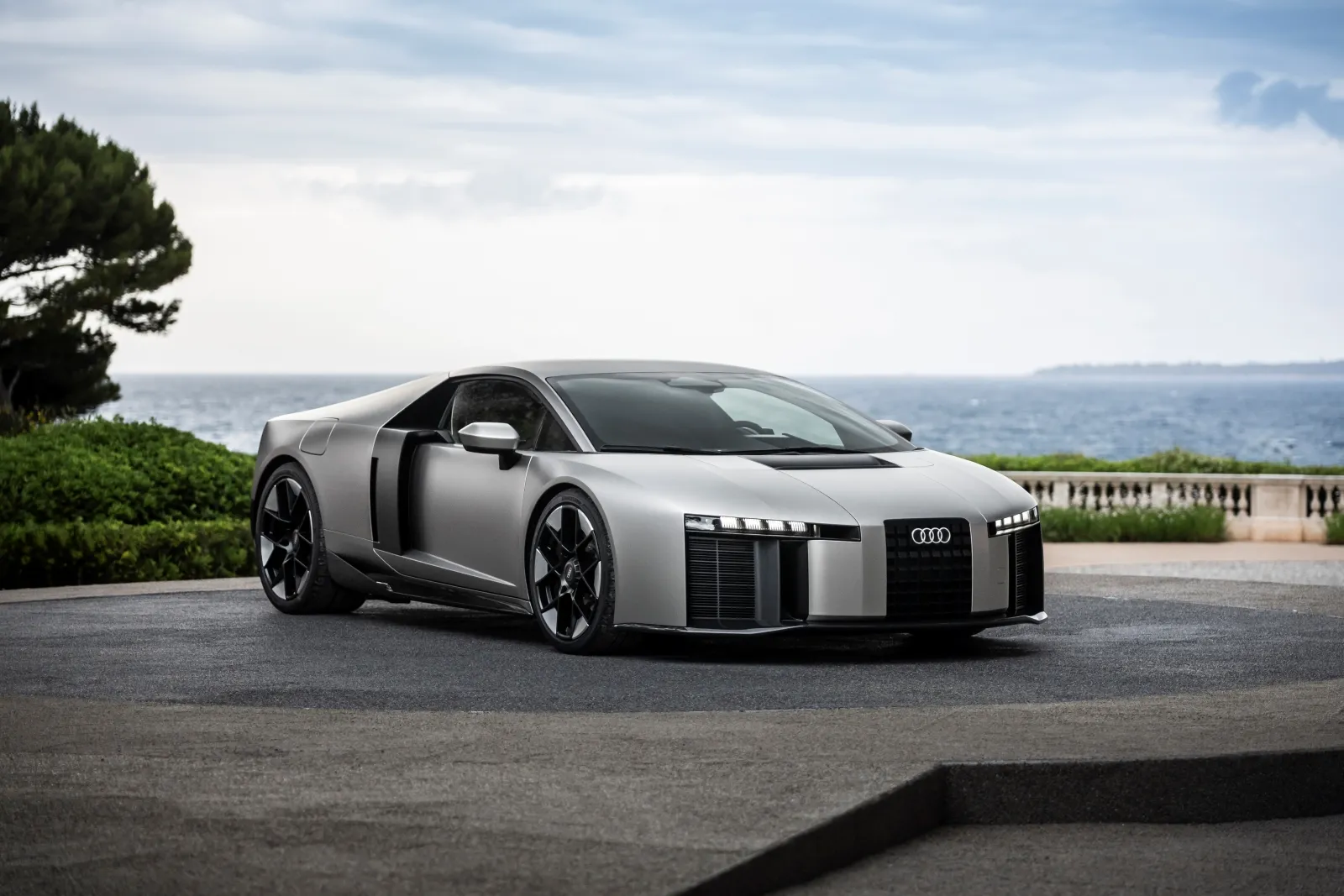 A sleek, silver sports car with a modern design is parked near a scenic coastal area, featuring a clear blue sky and lush greenery in the background.