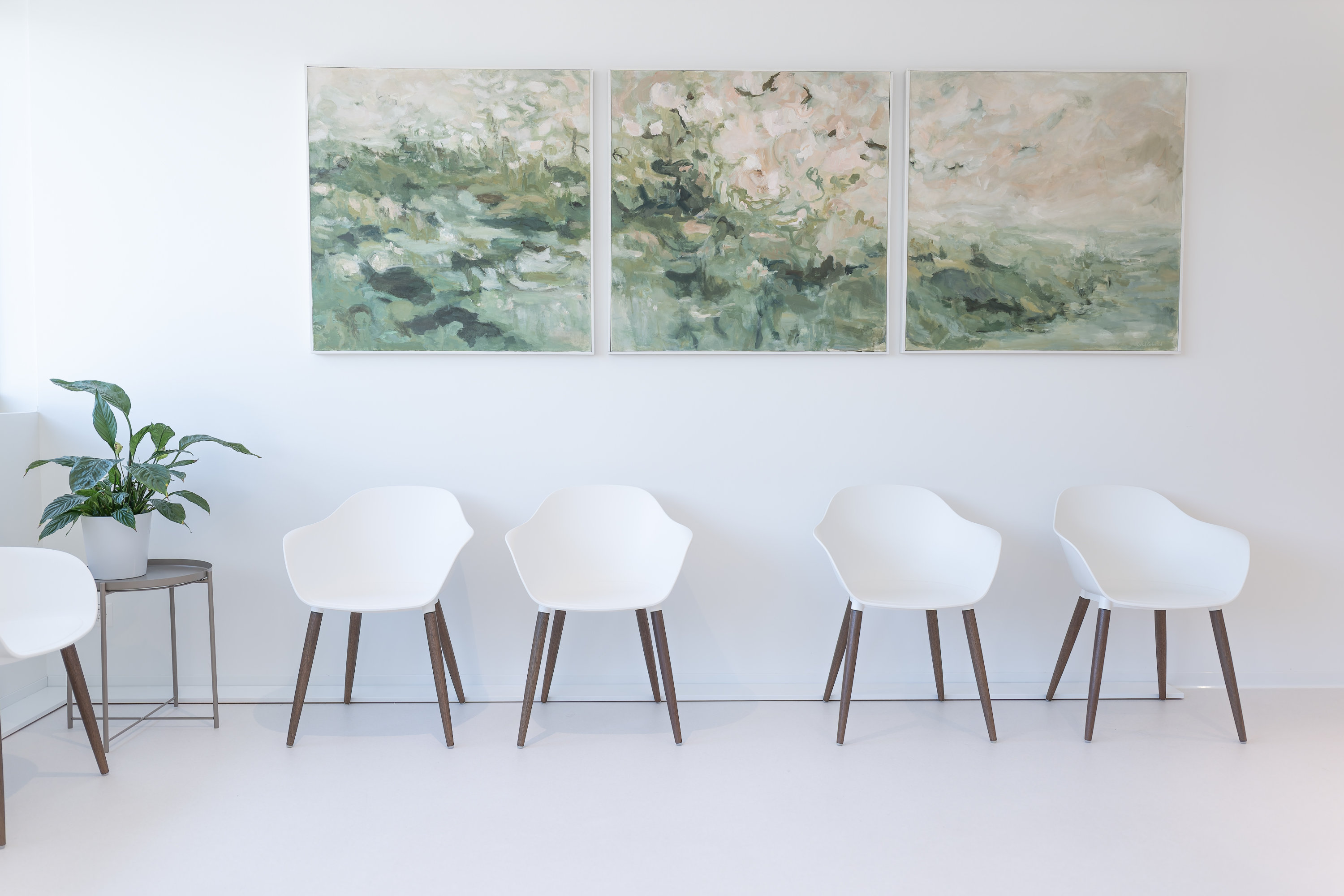 Eine Reihe von vier weißen Stühlen mit Holzbeinen säumt eine weiße Wand im Handzentrum Pratteln. Auf einem kleinen Tisch links steht eine Topfpflanze, und über den Stühlen hängen drei abstrakte grün-weiße Bilder.