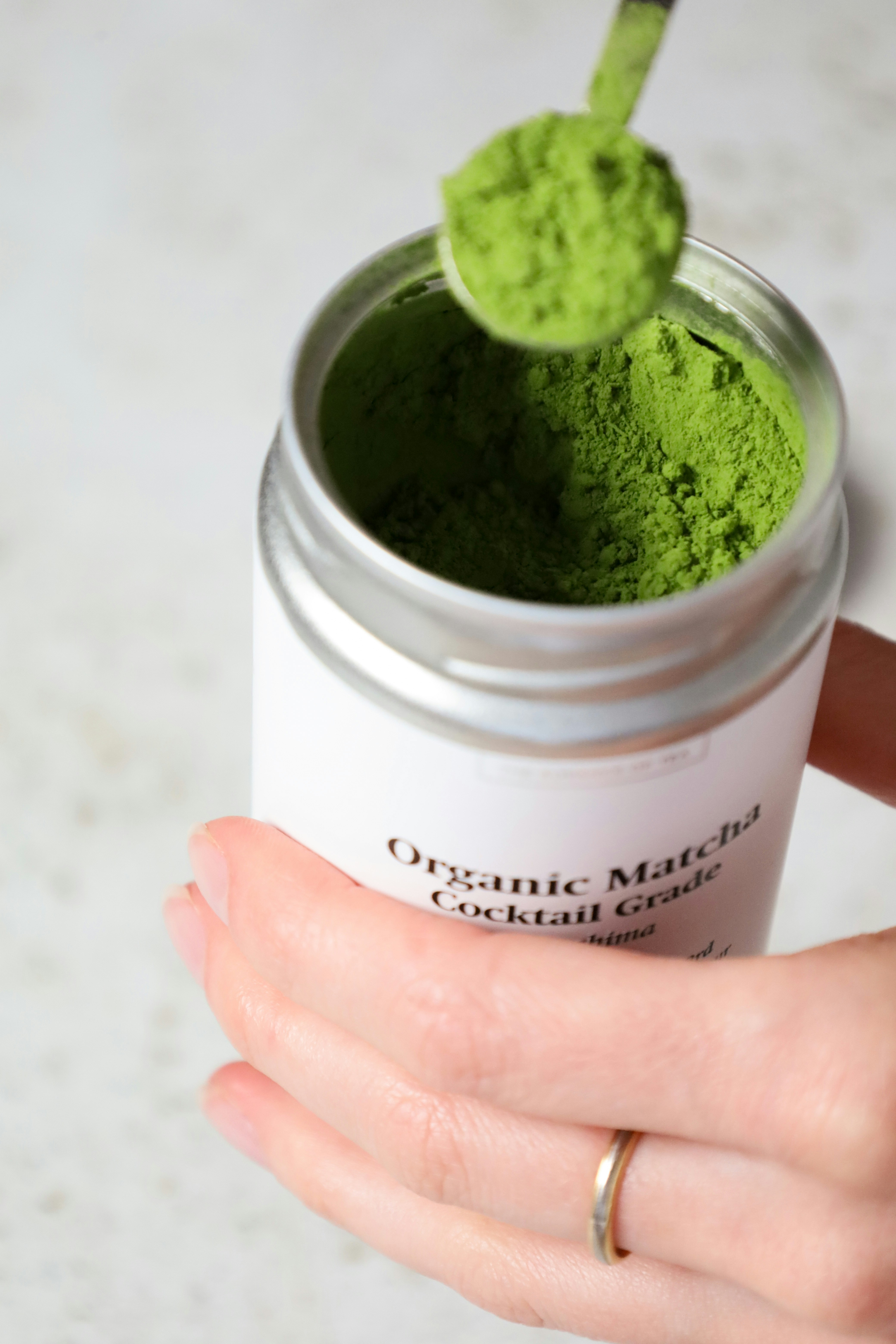 A hand holding a jar filled with matcha powder, a scoop partially inside the jar.
